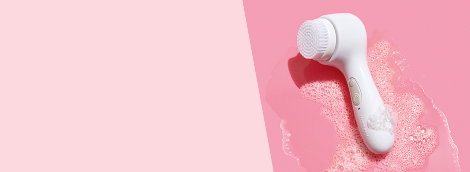 Die Bürste des Mary Kay Skinvigorate Sonic® Hautpflegesystems liegt auf einem pinken Hintergrund umgeben von feinem Seifenschaum.
