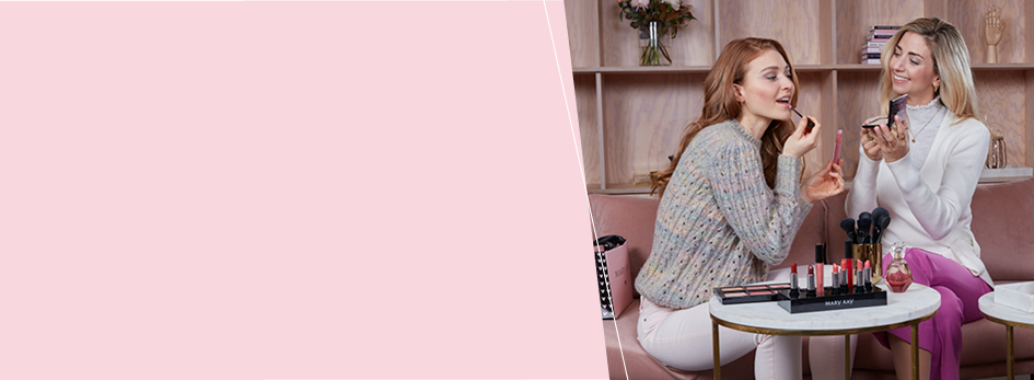 Eine selbständige Schönheits-Consultant mit Mary Kay berät eine Kundin. Sie sitzen auf einer rosa Couch, die Consultant hält einen kleinen Spiegel in der Hand und die Kundin testet einen Lipgloss. Auf einem kleinen Tisch stehen weitere Mary Kay Produkte. 