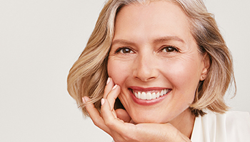 Eine blonde Frau mit reifer Haut stützt ihr Kinn in ihre Hand und lächelt in die Kamera