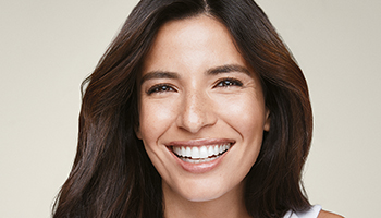 Eine Frau in den 30ern mit dunklen, langen Haaren und strahlender, reiner Haut lächelt frontal in die Kamera