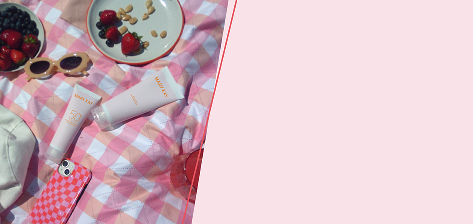 Eine rosa weiß karierte Picknickdecke von oben fotografiert. Man sieht die Mary Kay Sunscreen 50+ Sonnencreme, die Mary Kay After Sun Lotion sowie ein Handy, eine Sonnenbrille, ein Getränk, Beeren und einen Sonnenhut.