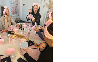 Mehrere Frauen mit einem rosa Stirnband in den Haare sitzen während einer Mary Kay Party mit einer selbständigen Schönheits-Consultant an einem Tisch, auf dem einige Mary Kay Produkte, Spiegel etc. zu sehen sind.